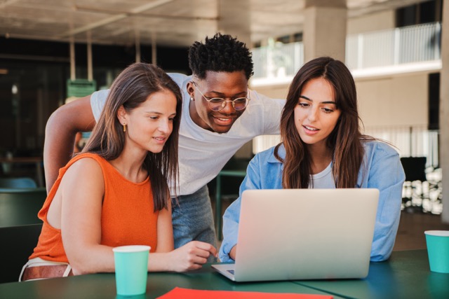 Three students sitting outdoors, collaborating around a laptop.